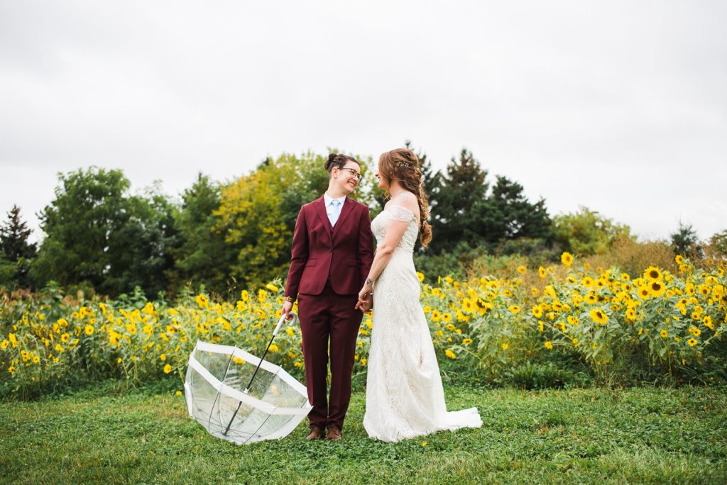 Two women on their wedding day holding umbrella in sunflower feild