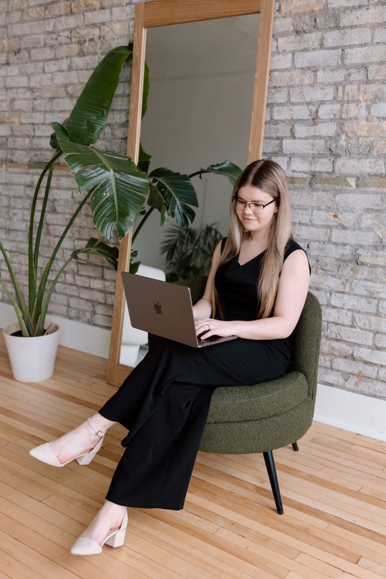 Katelyn in a black pantsuit sitting on a green chair typing on a laptop