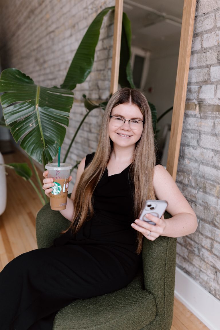Katelyn in a black pantsuit sitting on a green chair with iced coffee and phone in hand